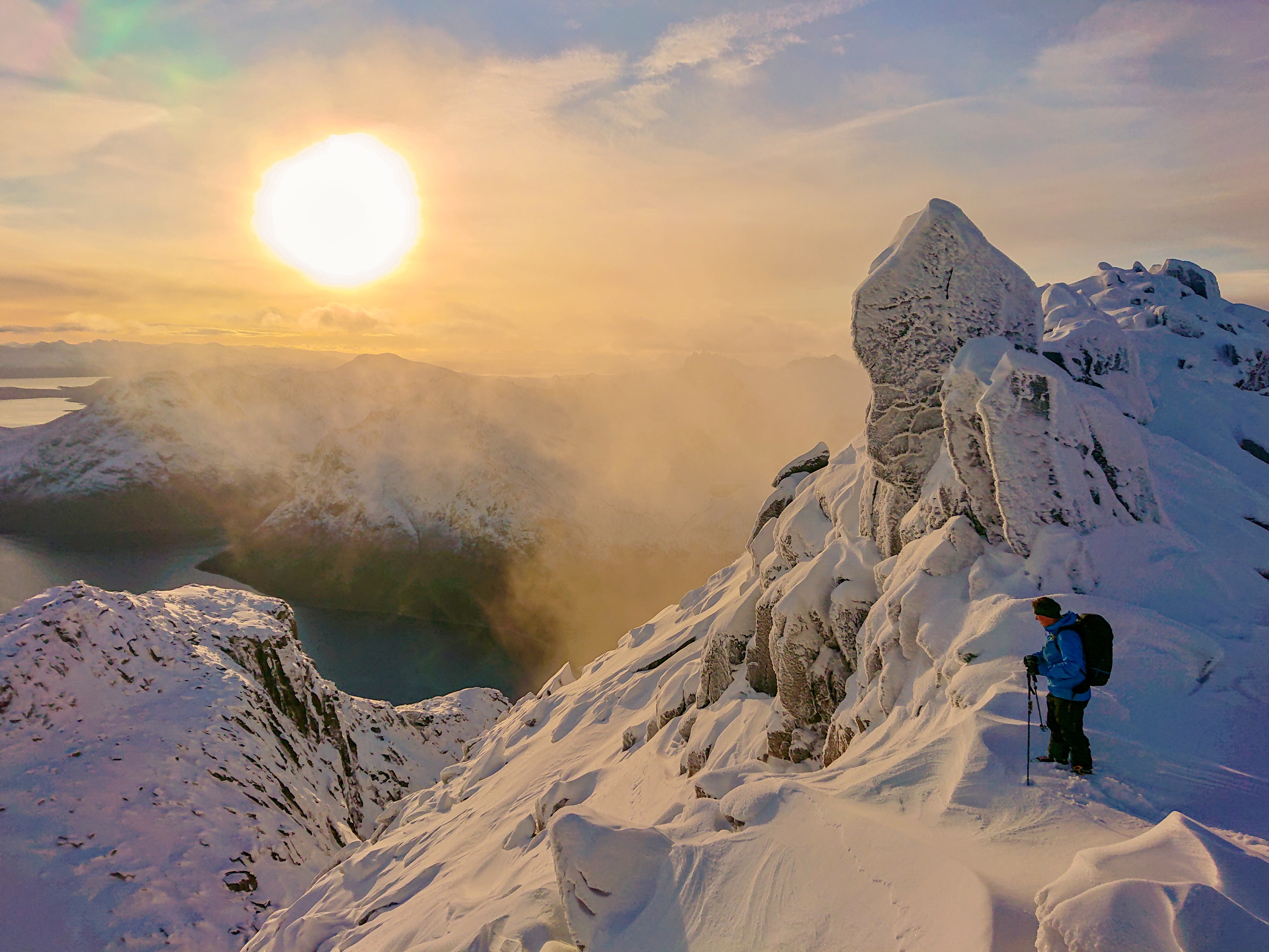A skier in a blue jacket stands on a snow-rimed ridge above a fjord as low sun breaks through drifting cloud